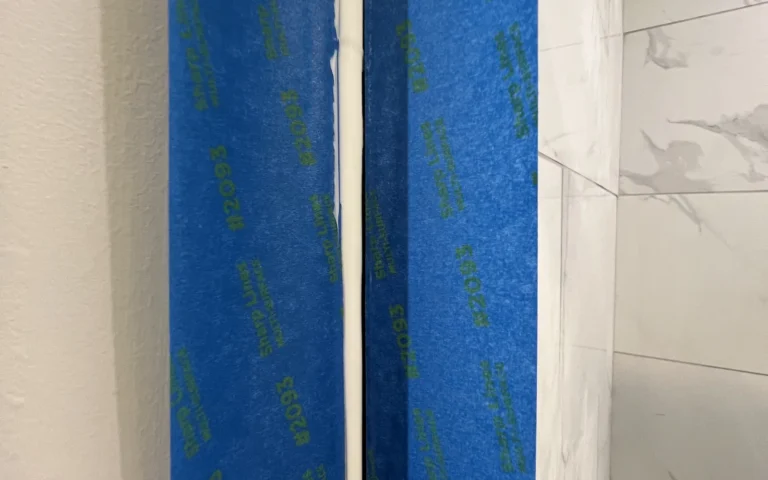 Blue painters tape and fresh white caulk sealing the gap between a wall and shower tiles