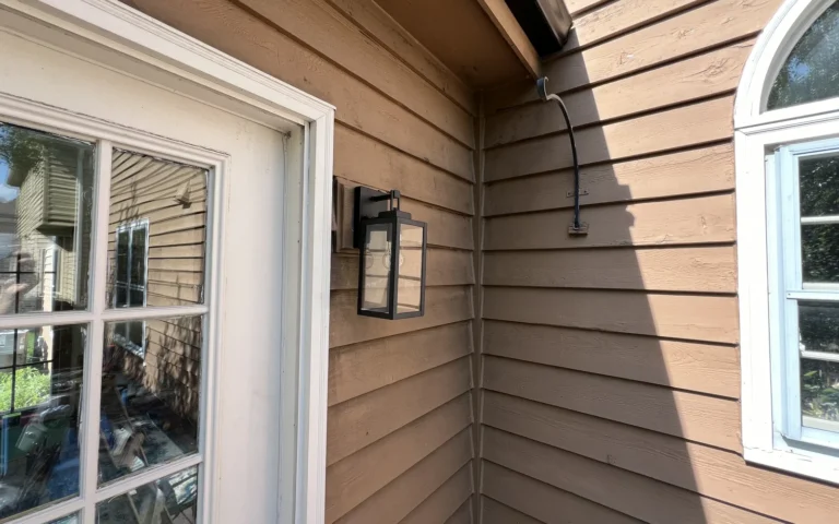 Black outdoor wall light fixture installed on brown siding near a white door