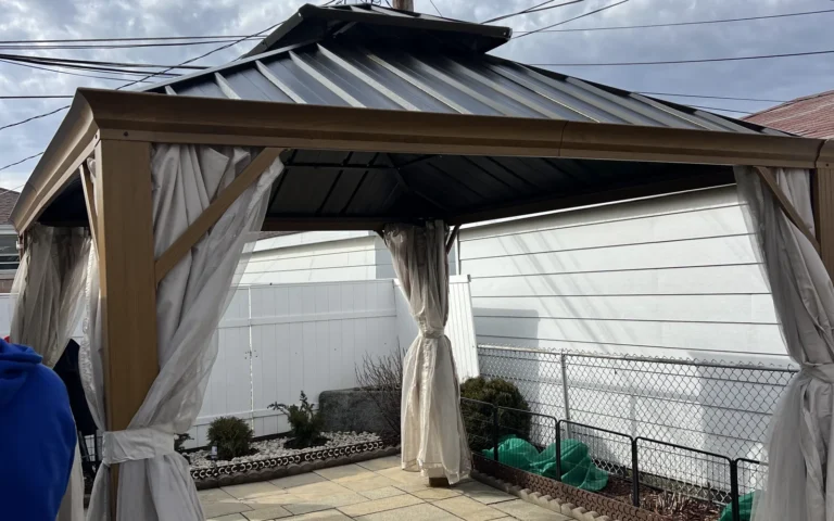 Completed wooden gazebo with metal roof and white curtains on a stone patio