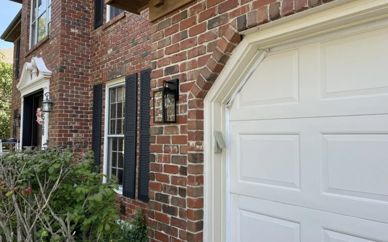 Black outdoor wall light fixture installed on brick house exterior near garage door
