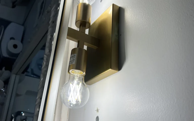 Gold bathroom wall sconce light fixture installed next to mirror with lit bulbs